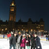 A group of people smile at a camera while skating in a rink in front of Parliament.