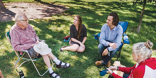A group of people of various ages sit in a circle in lawn chairs and on the grass, surrounded by trees.