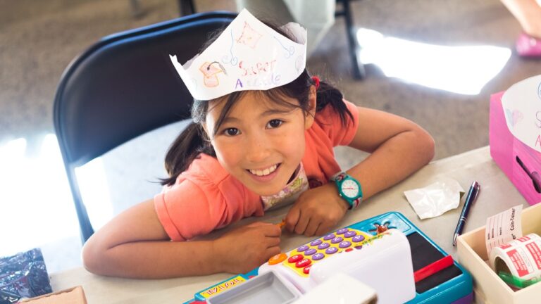 Photo of young girl in a handmade paper hat seated at a table with a toy cash register.