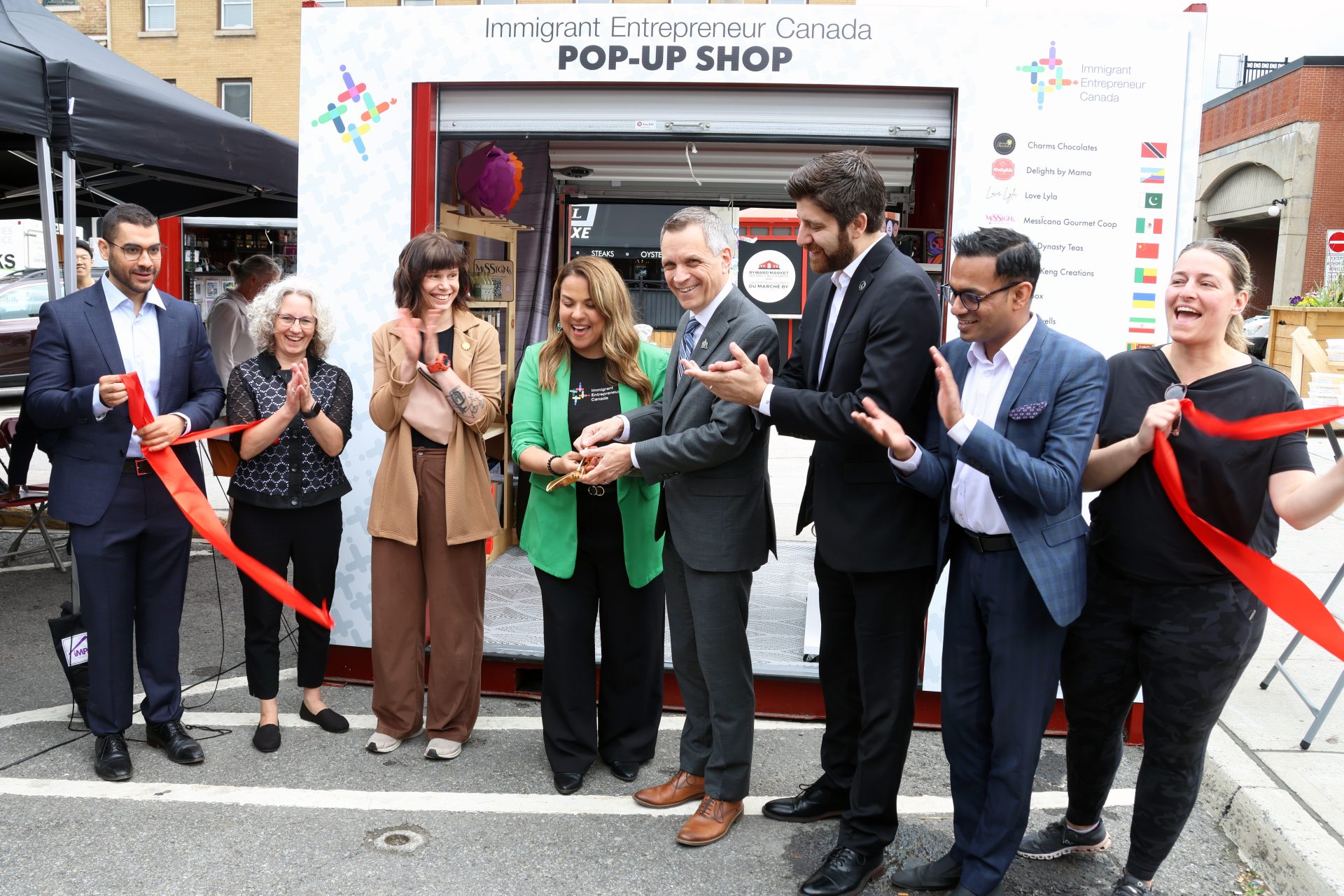 From left, Yasser Ghazi (Meridian), Paula Hopkins (Invest Ottawa), Anastasia Temborska (owner of PINBOX), Immigrant Entrepreneur Canada founder and CEO Karla Briones, Ottawa Mayor Mark Sutcliffe, Syrian-Canadian entrepreneur Tareq Hadhad (Peace by Chocolate), Daniel Thiruganaratnapathy (RBC) and Ottawa city councillor Stéphanie Plante (Rideau-Vanier) at the ribbon-cutting ceremony for IEC's Summer Retail Incubator 2024 program, which includes a pop-up shop in the ByWard Market. Photo by Caroline Phillips