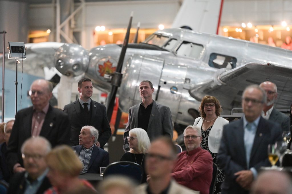 Left to right (standing): Trevor Friesen, (Edmonton Operations Manager, VHC), Wayne Duggan (Ottawa Operations Manager, VHC), and Amanda Mulawyshyn (Event organizer) join fellow attendees in watching the formal program during Veterans’ House Canada’s Andy Carswell Building 5th Anniversary celebration at the Canadian Aviation and Space Museum.