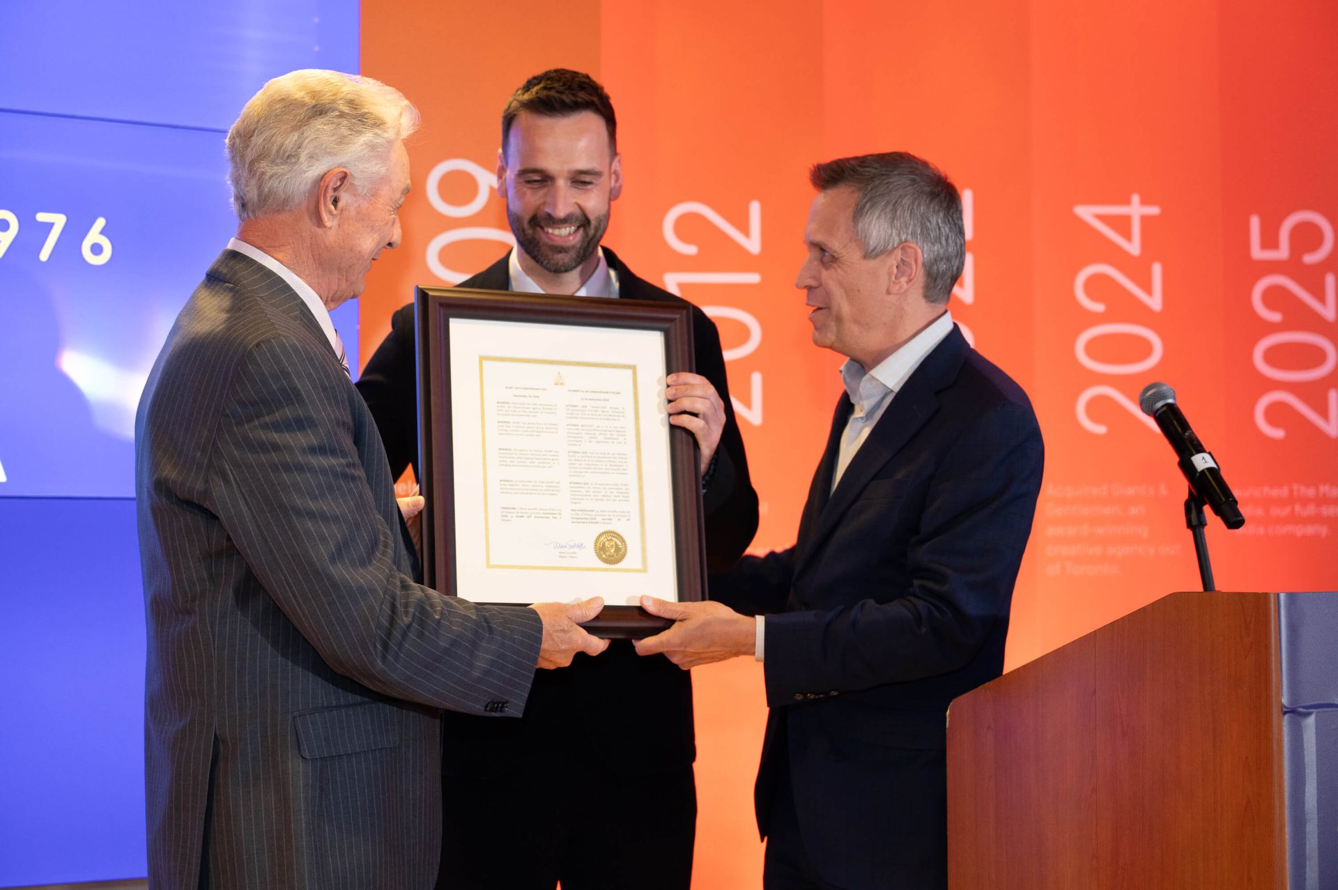 Mayor Mark Sutcliffe presenting Al Albania (left) and Andrew McWiggan of Acart Communications with an official mayoral proclamation at the Acart 50th anniversary party on Sept. 24, 2026 at the Westin Ottawa&rsquo;s TwentyTwo. Photo by Mark Holleron for OBJ.