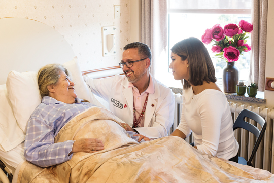 A doctor and young woman sit at the bedside of an elderly woman, who is smiling.